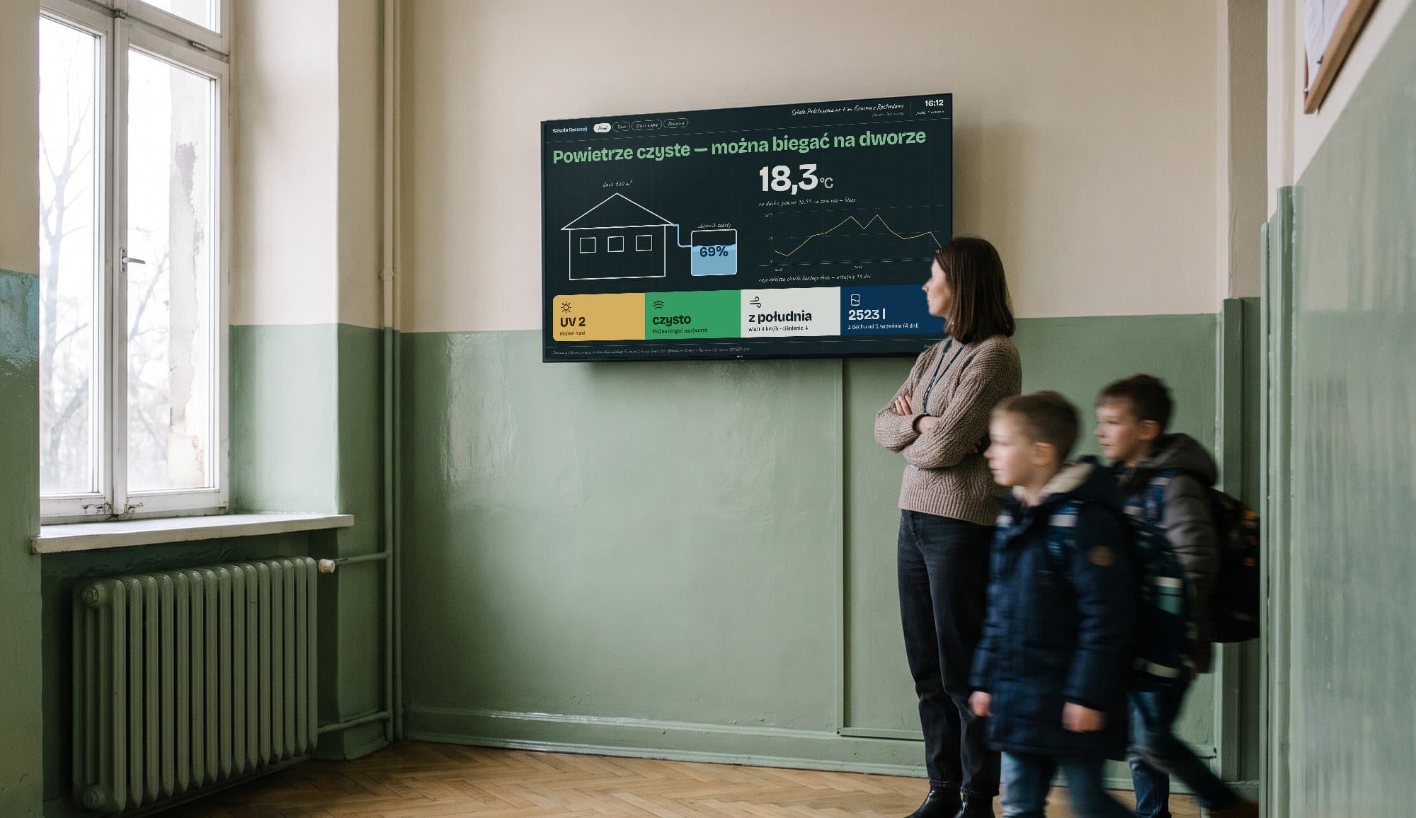 Monitor Szkoły Retencji na ścianie szkolnego korytarza; nauczycielka patrzy na ekran, obok przechodzą uczniowie.